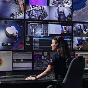 Operator monitors multiple security camera feeds on a wall of screens in a control room.
